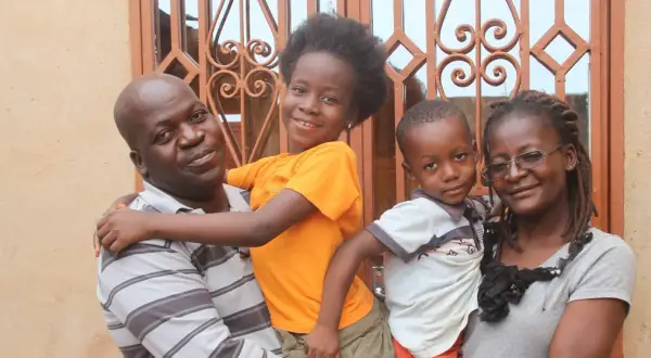 Famille souriante devant une fenêtre ornée. L'homme porte un enfant, la fille étreint son père. L'origine d'Anthony Samama est peut-être liée à cette joie familiale.