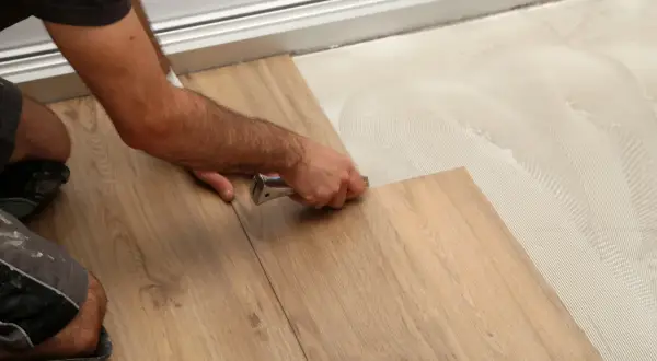 A person installs wood-look drop and lock flooring, using a tool to secure a plank onto adhesive-covered subfloor.