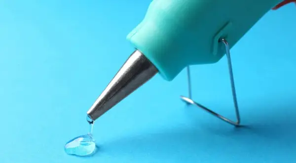 Un pistolet à colle turquoise dépose une goutte de colle transparente sur un fond bleu. Une petite tache de colle se forme.