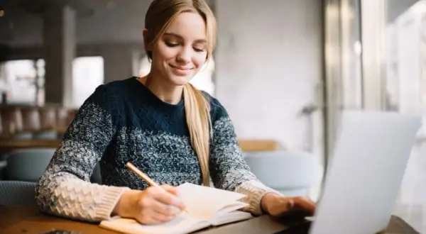 Uśmiechnięta studentka notuje w zeszycie, korzystając z laptopa. Cieszy się z możliwości, jakie dają jej zniżki studenckie.