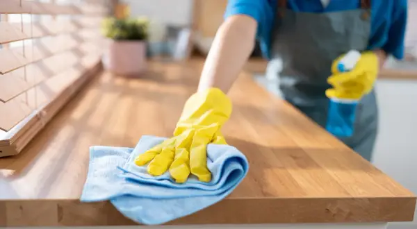 Ręka w żółtej rękawiczce z niebieską ściereczką czyści drewniany blat kuchenny. Dowiedz się, czym wyczyścić blat kuchenny laminowany, by lśnił czystością.