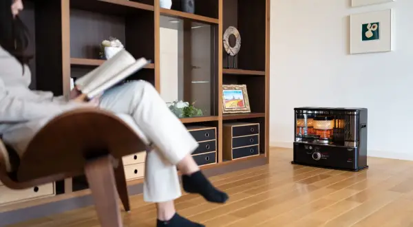 Donna legge un libro seduta su una poltrona, mentre un fungo a cherosene scalda l'ambiente. Una casa senza riscaldamento, ma con un tocco di calore.