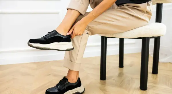 Kobieta w beżowych spodniach i czarnych sneakersach pokazuje, jak szybko wysuszyć buty.
