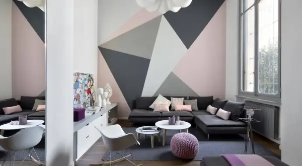 Soggiorno moderno con parete decorata con idee pittura geometrica in rosa, grigio e bianco. Divano angolare, tavolini e sedie di design.