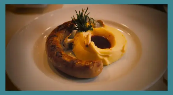 Une saucisse grillée, courbée sur une purée onctueuse, garnie de romarin et de sauce. Un plat réconfortant de saucisse purée.