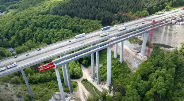Widok z lotu ptaka na budowę betonowego mostu nad lasem, z ruchem samochodowym na ukończonej części.
