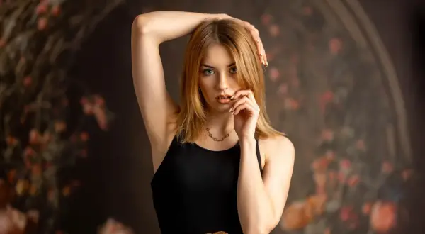 Zmysłowe zdjęcie modelki w czarnej sukience z złotym paskiem, z ręką przy twarzy.