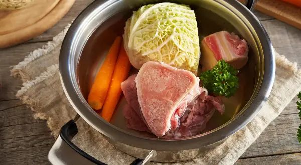 Une marmite de **potée au chou** avec des os de bœuf, des carottes, du chou et du persil, prête à mijoter.