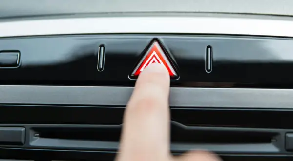 Un doigt appuie sur le bouton des feux de détresse obligatoires, un triangle rouge lumineux sur le tableau de bord d'une voiture.
