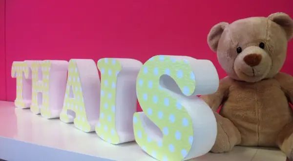 Lettre en relief "THAIS" décorée de pois sur fond rose vif, à côté d'un ours en peluche.