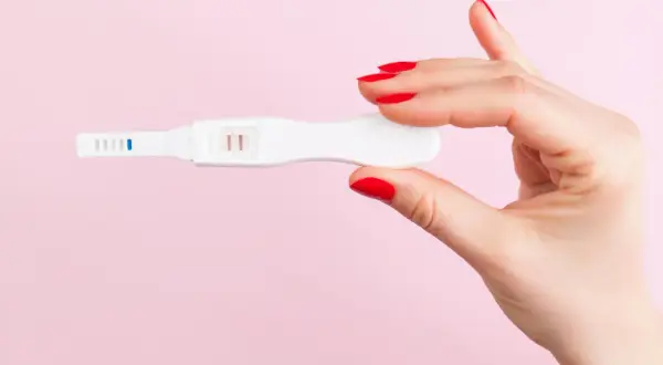 Dłoń z czerwonymi paznokciami trzyma test ciążowy z dwoma kreskami, sugerującymi ciążę biochemiczną.