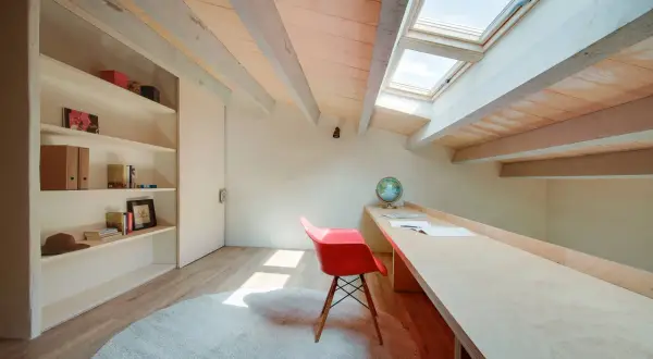 Un pequeño patio de luz interior baña con sol un escritorio de madera, una silla roja y una estantería.