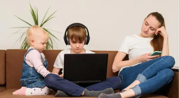 Dzieci na kanapie, chłopiec w słuchawkach gra na laptopie, maluch obok. Matka z telefonem. Kontrola rodzicielska w Polsce trwa do 18 lat.