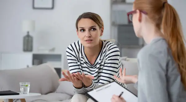 Kobieta w pasiastej bluzce gestykuluje, opowiadając o swoich problemach. Terapeuta z notatkami słucha uważnie, budując relację terapeutyczną.