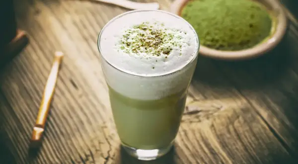Szklanka z zielonym matcha latte z pianką, obok miseczka z proszkiem matcha i bambusowa trzepaczka.