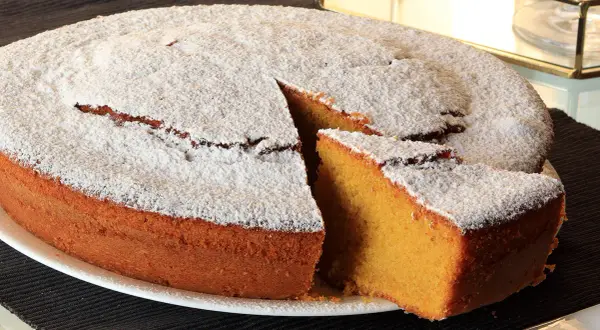 Una soffice torta carote e mandorle, cosparsa di zucchero a velo, con una fetta tagliata che ne rivela l'interno dorato.
