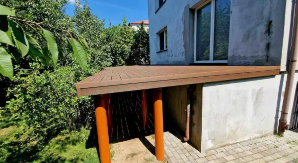 Nowy taras na słupach betonowych, z brązową, drewnopodobną nawierzchnią, otoczony zielenią.