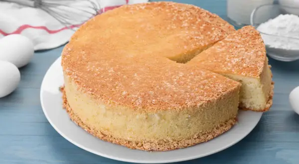 Puszysty biszkopt na talerzu, obok jajka, mąka, mleko i trzepaczka.