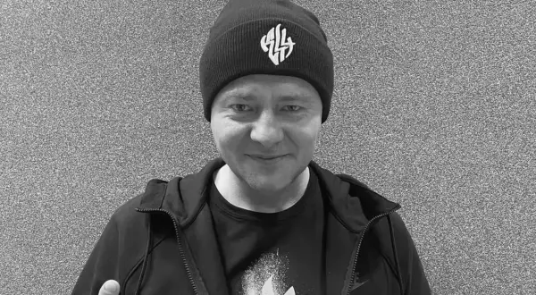Mężczyzna w czapce z logo, w bluzie i koszulce. W tle szara ściana. Nie wiadomo, **joka na co zmarł**.