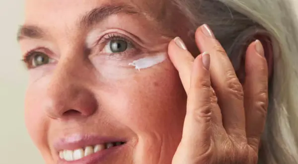 Eine Frau um die 60 trägt Augencreme auf. Ihre Haut zeigt natürliche Spuren des Alters, aber keine Pickel.