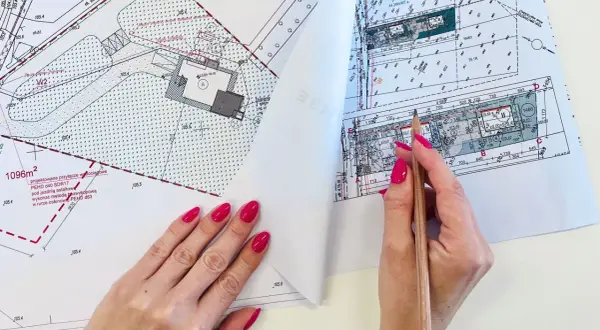 Ręce z malowanymi paznokciami przeglądają mapę do celów projektowych, wskazując ołówkiem szczegóły planu zagospodarowania terenu.