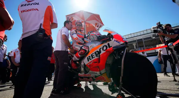 Piloto de MotoGP Repsol Honda listo en la parrilla de salida, bajo un sol radiante y rodeado de su equipo y cámaras.