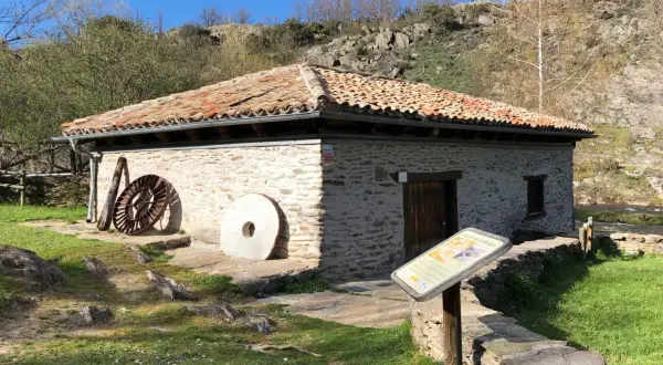 El molino de la Hiruela, con su tejado de tejas rojas y muros de piedra, se alza junto a un río. A su lado, piedras de moler y una rueda de hierro evocan su pasado.