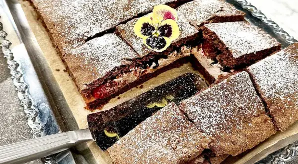 En chokladgrädde tårta skuren i bitar, pudrad med florsocker och dekorerad med violer. En tårtspade lyfter upp en bit.