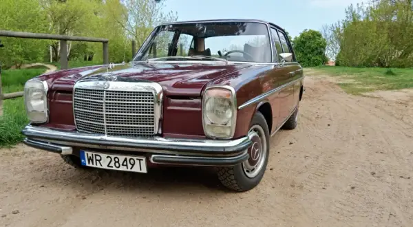 Klasyczny mercedes w114 w bordowym kolorze, zaparkowany na piaszczystej drodze wśród zieleni.