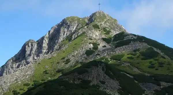 Widok na szczyt Giewontu z krzyżem, zielone zbocza i kamieniste ścieżki. W dole grupa turystów rozpoczyna wejście na Giewont.