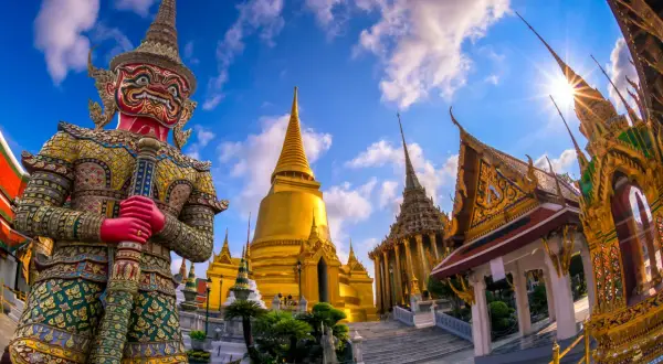 Imponerande tempel i Bangkok med en stor, färgglad jättefigur framför gyllene pagoder under en blå himmel med moln.