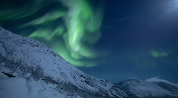 Grüne Nordlichter tanzen über schneebedeckte Berge bei Tromsø. Ein magischer Moment, der zu den Tromsø Geheimtipps gehört.