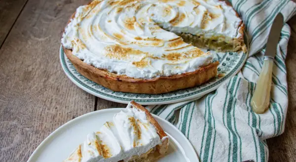 Une part de tarte rhubarbe meringuée, avec sa meringue dorée et sa garniture acidulée, sur une assiette blanche.