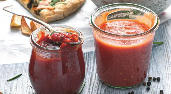 Dwa słoiki domowego sosu do pizzy, jeden z łyżką pełną aromatycznej masy. Obok kawałek pizzy i przyprawy.