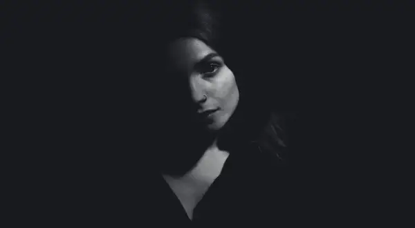 A woman's face emerges from darkness, her features dramatically sculpted by side lighting. A subtle nose ring catches the light.