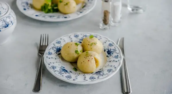 Trzy pyzy ziemniaczane na talerzu z niebieskim wzorem, udekorowane natką pietruszki. Obok widelec i nóż.