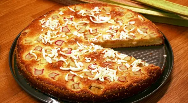 En saftig rabarberkladdkaka med smulad vit choklad och färska rabarberstjälkar i bakgrunden.