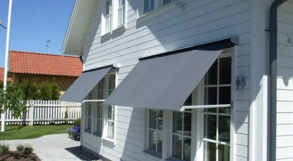 Grå markiser monterade på ett vitt trähus ger skugga åt fönstren. En vacker trädgård med stenläggning och staket syns i bakgrunden.