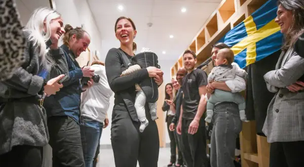 Elin Johansson, med ett leende och en bebis i bärsele, omgiven av glada människor och en svensk flagga. Kanske en paus från taekwondo-träningen?