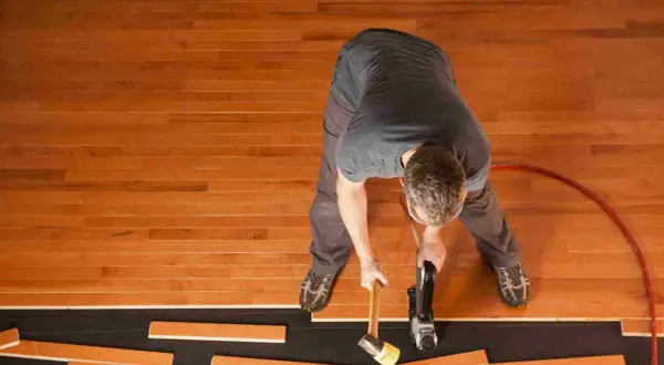 A man is installing waterproof flooring, using a nail gun and mallet to secure wooden planks.