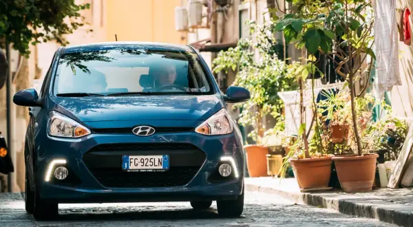 Blauer Hyundai i10 fährt auf einer Kopfsteinpflasterstraße, umgeben von Pflanzen und Gebäuden.