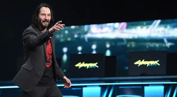 Keanu Reeves na scenie, wskazując palcem, w koszulce z logo Cyberpunk 2077. Gra zarobiła miliony, a on jest jej twarzą.