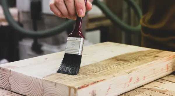 Mano che applica vernice trasparente su un pezzo di legno grezzo con un pennello. Guida su come verniciare il legno per proteggerlo.