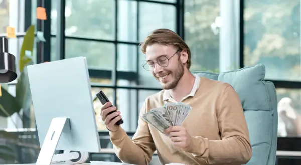 Uśmiechnięty mężczyzna z okularami trzyma telefon i pieniądze. Zastanawia się, co wpisać w tytule przelewu dla kolegi.
