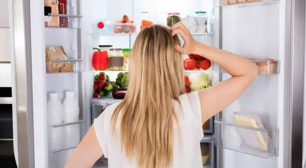 Donna confusa davanti al frigo pieno di cibo, con il desiderio di mangiare e non ingrassare, ma lo stress la blocca.