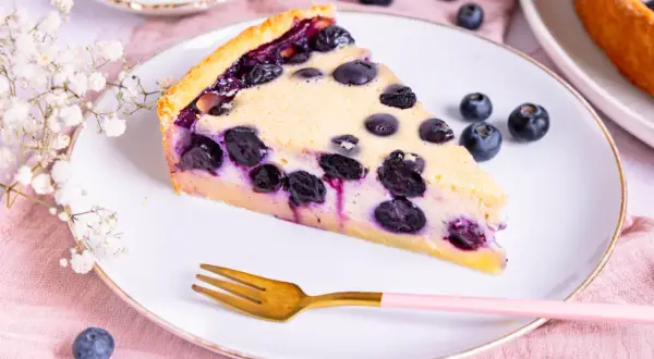 Ein Stück Heidelbeerkuchen mit Vanillepudding, garniert mit frischen Heidelbeeren und einer zarten Blumen-Deko.