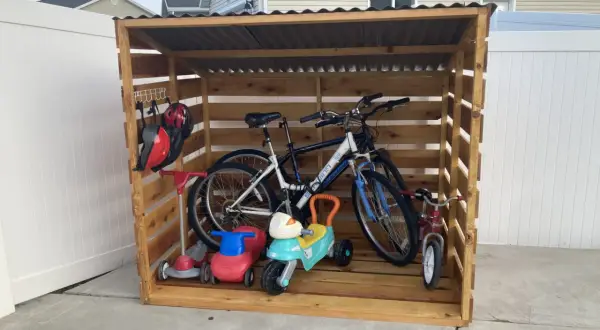 A DIY bike rack made of wood stores two adult bikes, a tricycle, and two ride-on toys. Helmets hang on a hook.