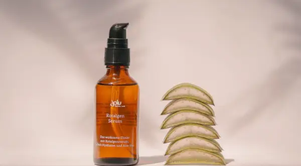 Braunes Fläschchen mit Rotalgen Serum, das Rotalgenextrakt als geliermittel enthält, neben aufgestapelten Aloe-Vera-Scheiben.