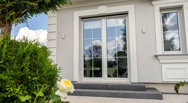 Eleganckie okno bez klamki z białymi szprosami, odbijające błękitne niebo i chmury. Obok zielone krzewy i biała róża.