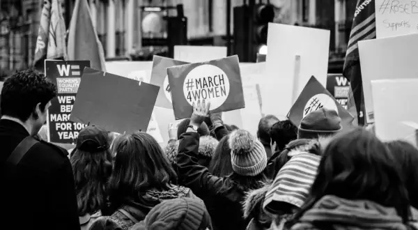 Tłum na marszu, gdzie feminizm i emancypacja idą w parze. Ludzie niosą transparenty, jeden z nich głosi "#MARCH 4WOMEN".